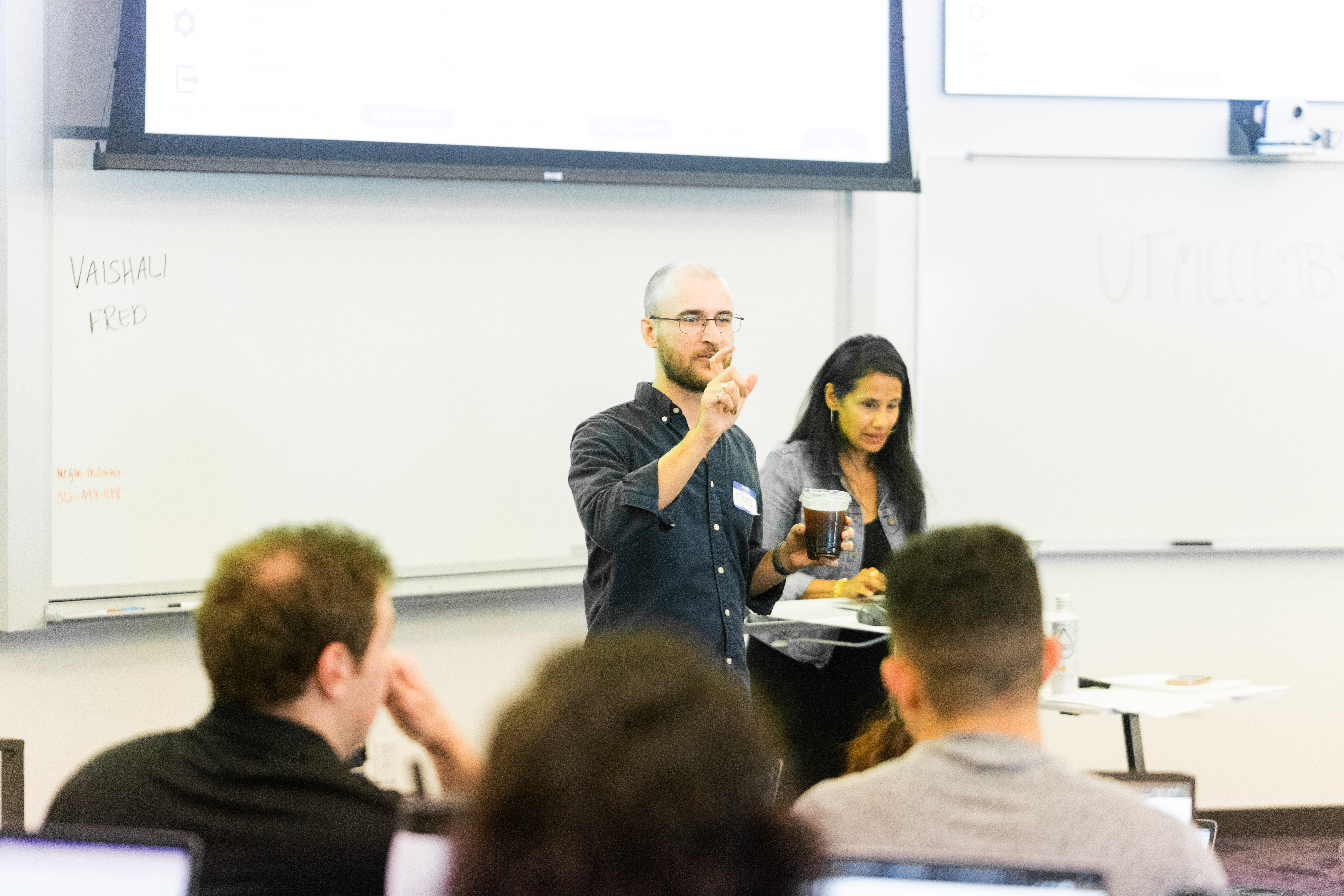 A presenter, Fred Tally-Foos, gesturing with one hand while holding a drink, stands at the front of a classroom with a woman beside him. Students are visible in the foreground, seated at desks with laptops.
